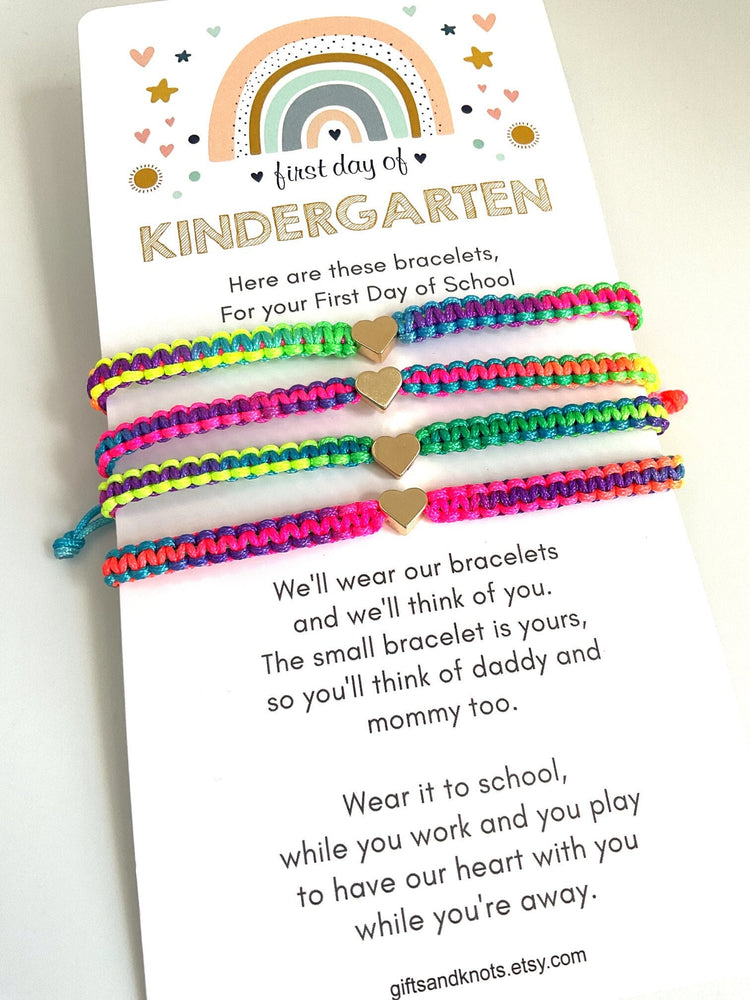 Rainbow Card First Day of KINDERGARTEN. The card has a message that reads 'Here are these bracelets, For your First Day of School. We'll wear our bracelets and we'll think of you. The small bracelet is yours, so you'll think of daddy and mommy too. Wear it to school, while you work and you play to have our heart with you while you're away. The card is decorated with small hearts and stars. There are four colorful bracelets with gold heart charms on the card.