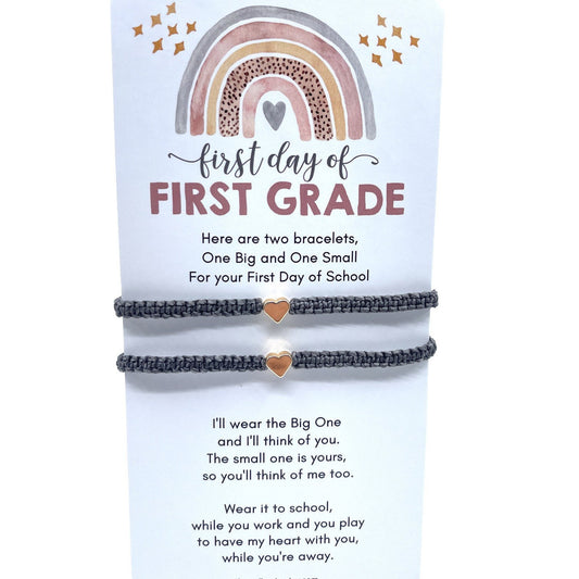 Two gray friendship bracelets with a small heart charm on each. The bracelets are displayed on a card with a rainbow design and the text 'first day of FIRST GRADE'. The card also includes the text 'Here are two bracelets, One Big and One Small For your First Day of School' and 'I'll wear the Big One and I'll think of you. The small one is yours, so you'll think of me too. Wear it to school, while you work and you play to have my heart with you, while you're aw