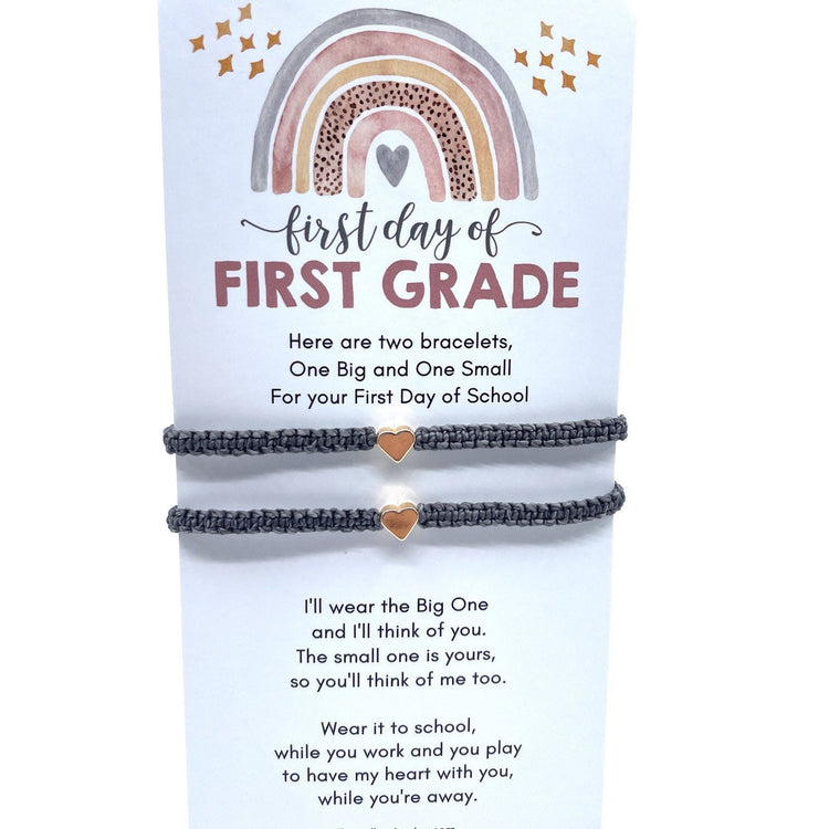 Two gray friendship bracelets with a small heart charm on each. The bracelets are displayed on a card with a rainbow design and the text 'first day of FIRST GRADE'. The card also includes the text 'Here are two bracelets, One Big and One Small For your First Day of School' and 'I'll wear the Big One and I'll think of you. The small one is yours, so you'll think of me too. Wear it to school, while you work and you play to have my heart with you, while you're aw