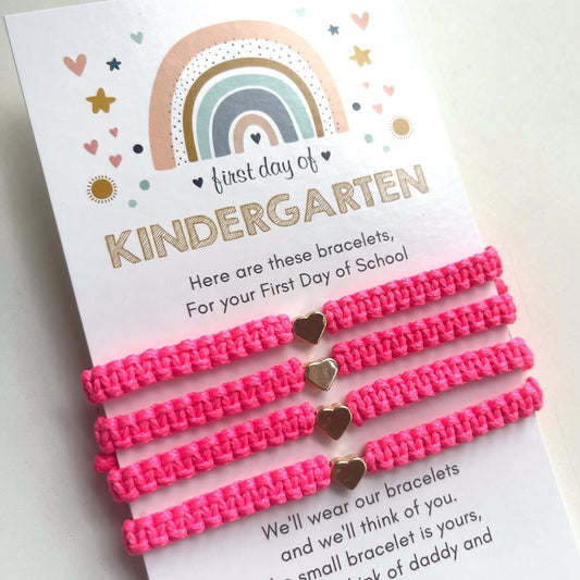Four neon pink friendship bracelets with gold heart charms on a white card with a rainbow design and a First day of KINDERGARTEN poem.