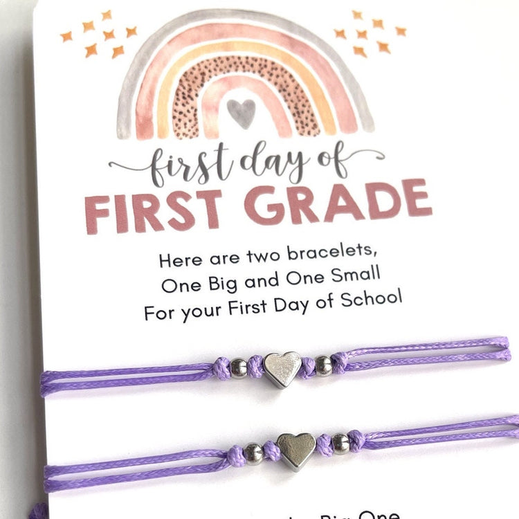 First day of FIRST GRADE card is accompanied by two purple friendship bracelets with silver heart charms. The text on the card reads 'Here are two bracelets, One Big and One Small For your First Day of School. I'll wear the Big One and I'll think of you. The small one is yours, so you'll think of me too. Wear it to school, while you work and you play to have my heart with you, while you're away.