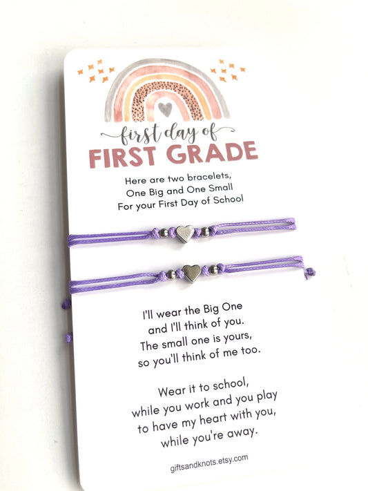 First day of FIRST GRADE card is accompanied by two purple friendship bracelets with silver heart charms. The text on the card reads 'Here are two bracelets, One Big and One Small For your First Day of School. I'll wear the Big One and I'll think of you. The small one is yours, so you'll think of me too. Wear it to school, while you work and you play to have my heart with you, while you're away.